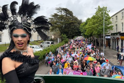 Drag queen Chae with a C, wearing a black sequinned gown and elaborate feathered headdress, stands on the upper deck of the Swansea Pride open-top bus looking back over the parade, with hundreds of marchers carrying flags and banners visible in the street behind her.