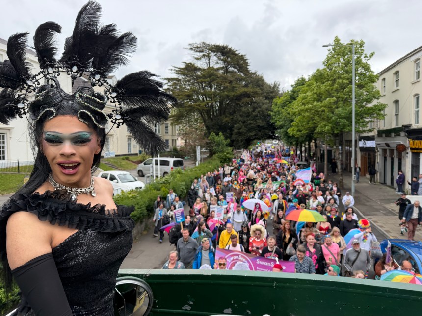 Drag queen Chae with a C, wearing a black sequinned gown and elaborate feathered headdress, stands on the upper deck of the Swansea Pride open-top bus looking back over the parade, with hundreds of marchers carrying flags and banners visible in the street behind her.