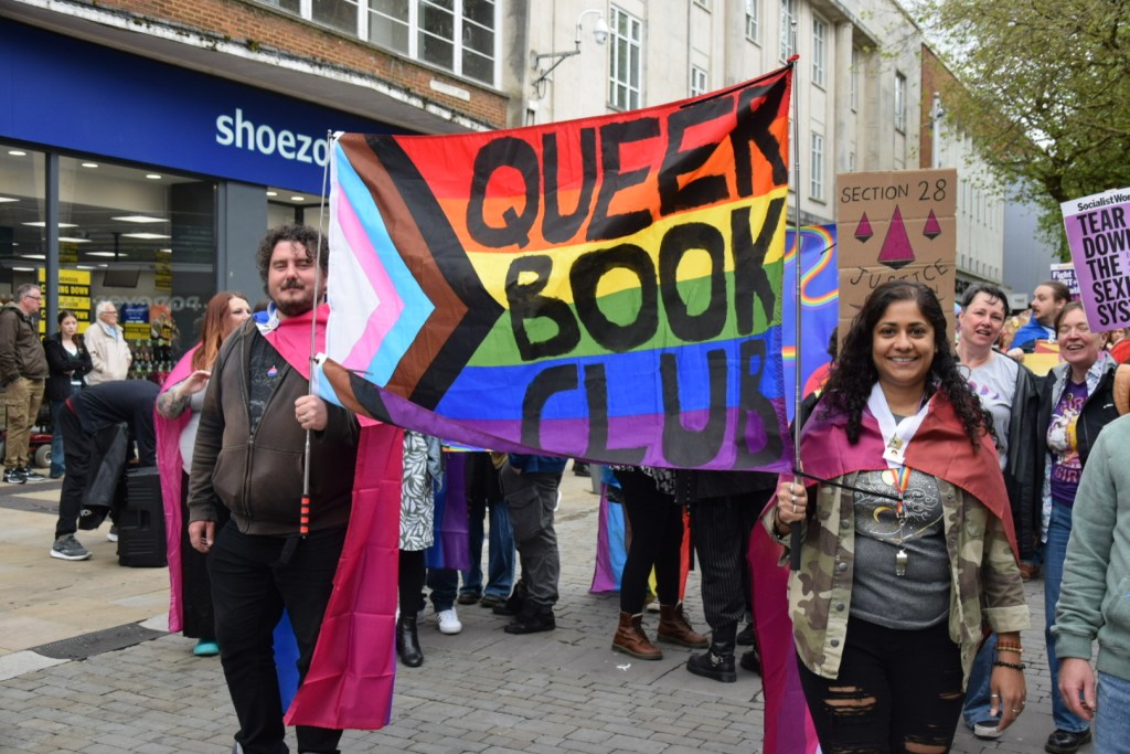 Two people carry a large colourful Queer Book Club banner decorated with the Progress Pride flag through Swansea city centre during the Pride 2026 parade, with other marchers and onlookers visible behind them.