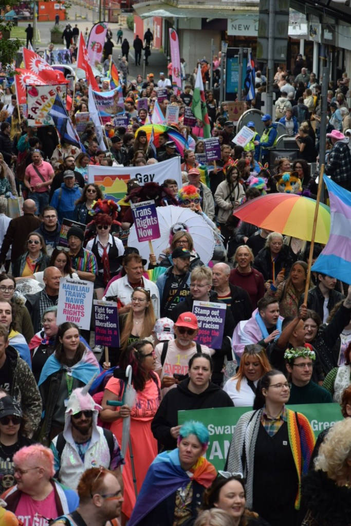 Overhead view of the Swansea Pride 2026 parade passing through the city centre, with the Quadrant Shopping Centre sign visible and hundreds of marchers carrying rainbow flags and Pride banners filling the street.