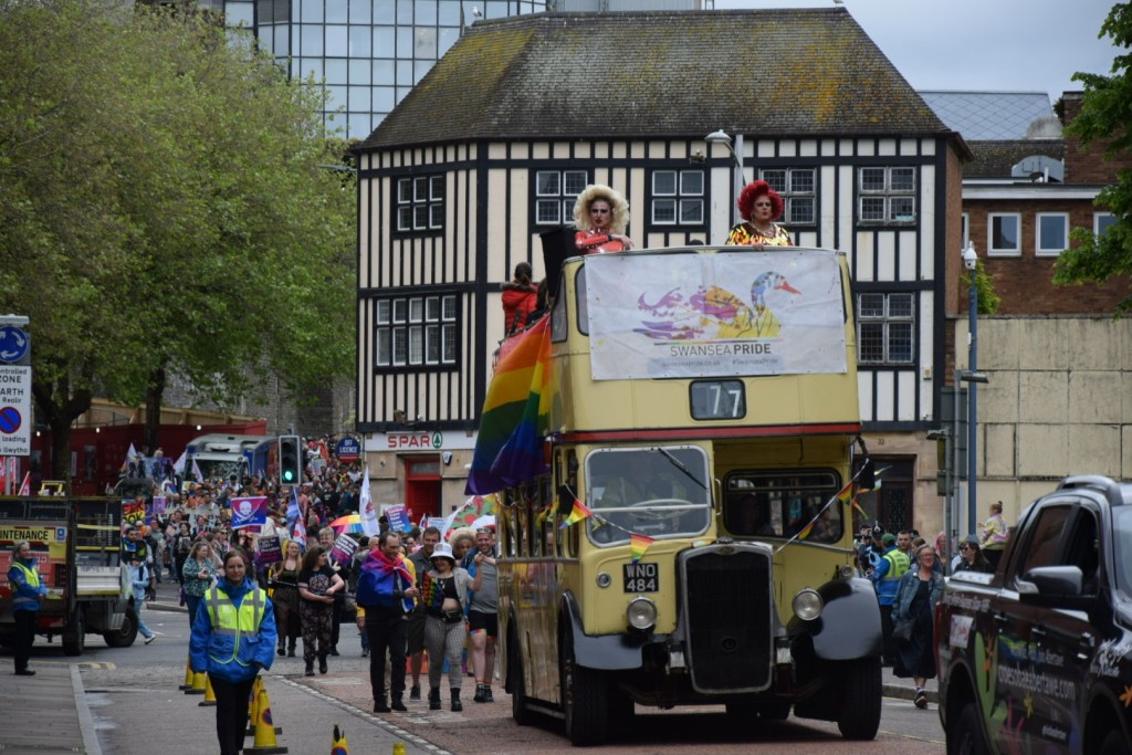 Drag queens Ruby Slippers and Diana D stand on the upper deck of a vintage yellow open-top double-decker bus bearing the Swansea Pride banner, with the parade route and a timber-framed Swansea building visible behind them.