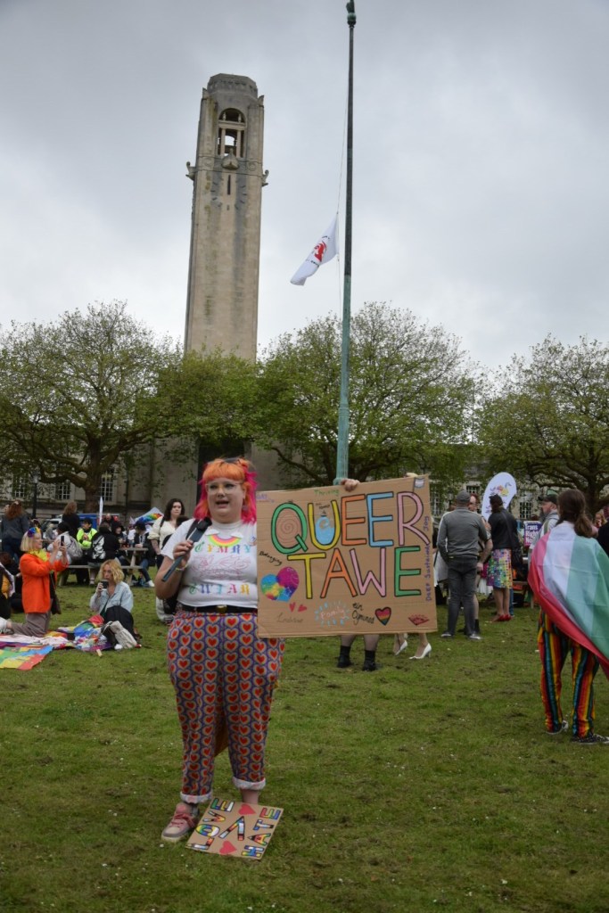 A woman with orange hair and colourful clothing holds a hand-painted Queer Tawe sign at the Swansea Pride 2026 festival outside the Guildhall, with the Grade I-listed Guildhall clock tower visible against an overcast sky behind her.