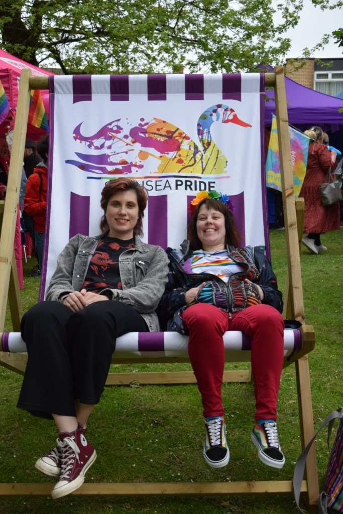Two smiling women sit in a giant Swansea Pride branded deckchair featuring the event's colourful swan logo, at the festival outside the Guildhall during Swansea Pride 2026.