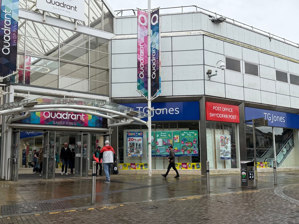 External view of the Quadrant Shopping Centre entrance in Swansea on a wet day, with branded Quadrant Shopping banners overhead, a TGJones store and Post Office counter visible to the right, and shoppers walking past.