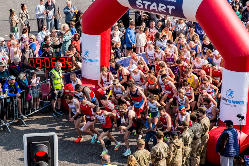View from above of the start line at a previous Mizuno Swansea Half Marathon, with runners in England, Wales and military team kit setting off through a red Front Runner Events arch, watched by crowds of spectators and Army personnel.