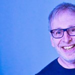 Comedian Ian Stone, a smiling man with short grey hair and dark-rimmed glasses, photographed against a blue background.