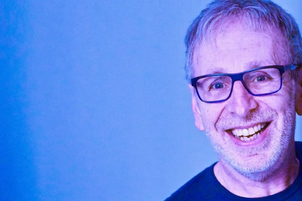 Comedian Ian Stone, a smiling man with short grey hair and dark-rimmed glasses, photographed against a blue background.