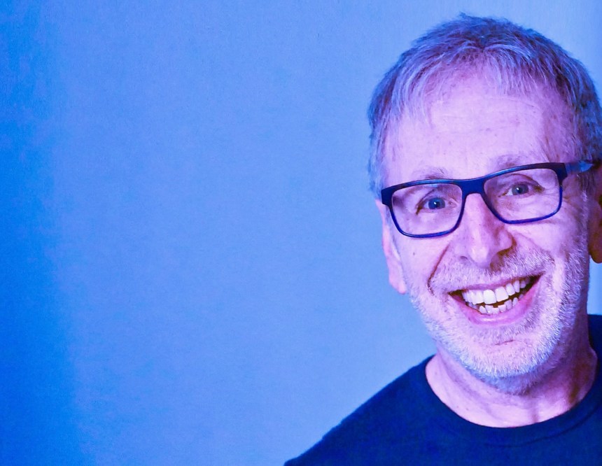 Comedian Ian Stone, a smiling man with short grey hair and dark-rimmed glasses, photographed against a blue background.