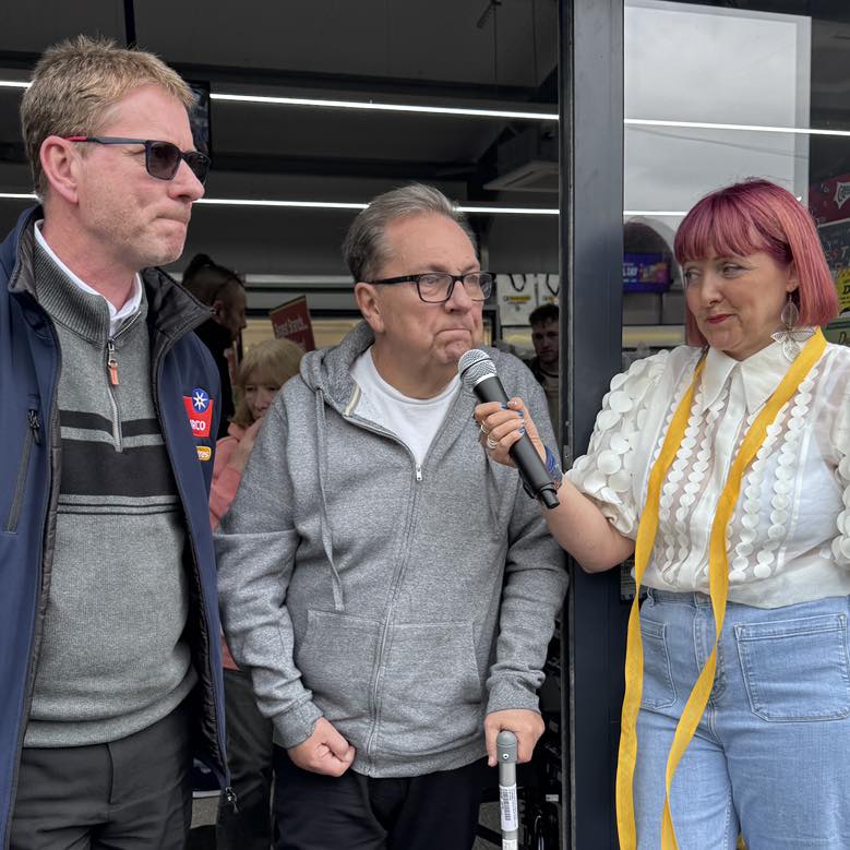 Kev Johns MBE holding a microphone and a walking stick at the entrance to CK Foodstores in Bynea, flanked by a man in a CK branded gilet and presenter Siany Martin with pink hair.