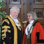 Two women in ceremonial civic robes and chains of office smiling together in front of ornate carved wooden doors decorated with climbing flowers — Cllr Penny Matthews in black and gold Lord Mayor's robes on the left and Cllr Susan Jones in red Deputy Lord Mayor's robes on the right.