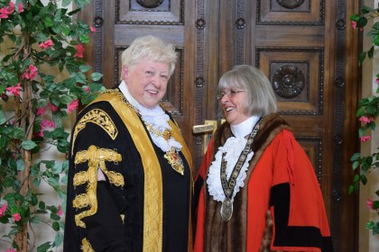 Two women in ceremonial civic robes and chains of office smiling together in front of ornate carved wooden doors decorated with climbing flowers — Cllr Penny Matthews in black and gold Lord Mayor's robes on the left and Cllr Susan Jones in red Deputy Lord Mayor's robes on the right.