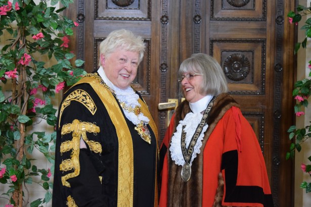 Two women in ceremonial civic robes and chains of office smiling together in front of ornate carved wooden doors decorated with climbing flowers — Cllr Penny Matthews in black and gold Lord Mayor's robes on the left and Cllr Susan Jones in red Deputy Lord Mayor's robes on the right.