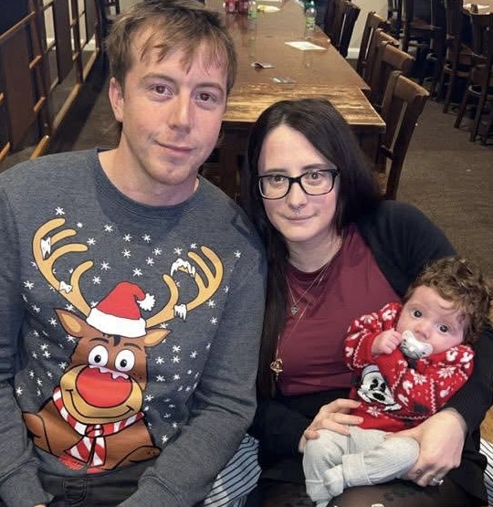 Liam John in a Christmas jumper and Shania Jenkins in a red top sit together at a table, with Shania holding a newborn baby Tobi dressed in a red Christmas outfit.
