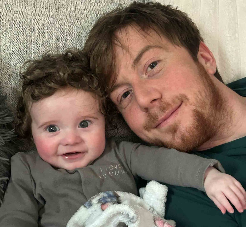 Liam John lying next to baby Tobi, who is wearing a grey babygrow reading "I Love My Mum" and smiling at the camera.