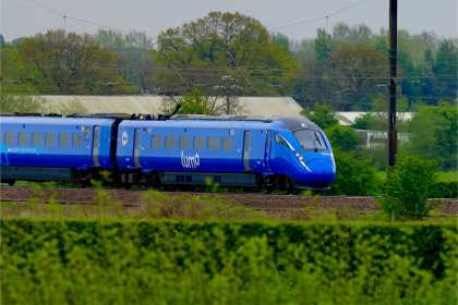 A blue Lumo high-speed train travelling along an electrified railway line through the countryside, with overhead line equipment, mature trees and a hedgerow in the foreground