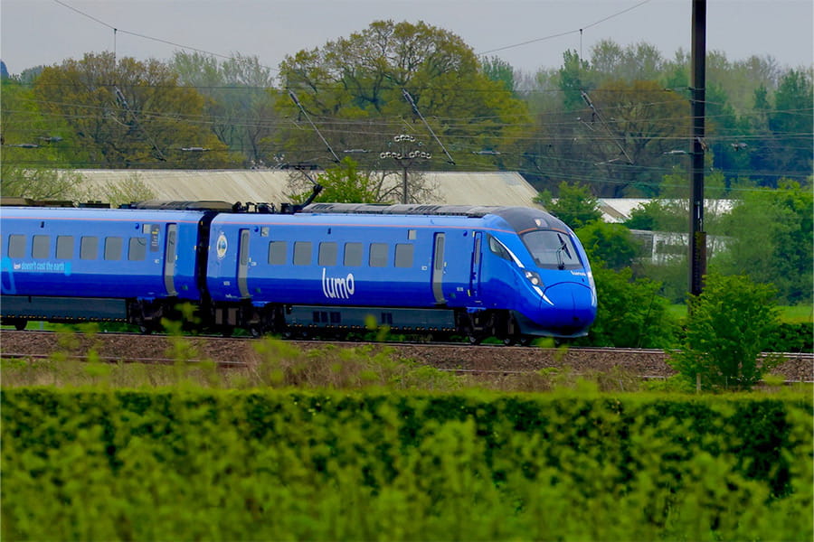 A blue Lumo high-speed train travelling along an electrified railway line through the countryside, with overhead line equipment, mature trees and a hedgerow in the foreground
