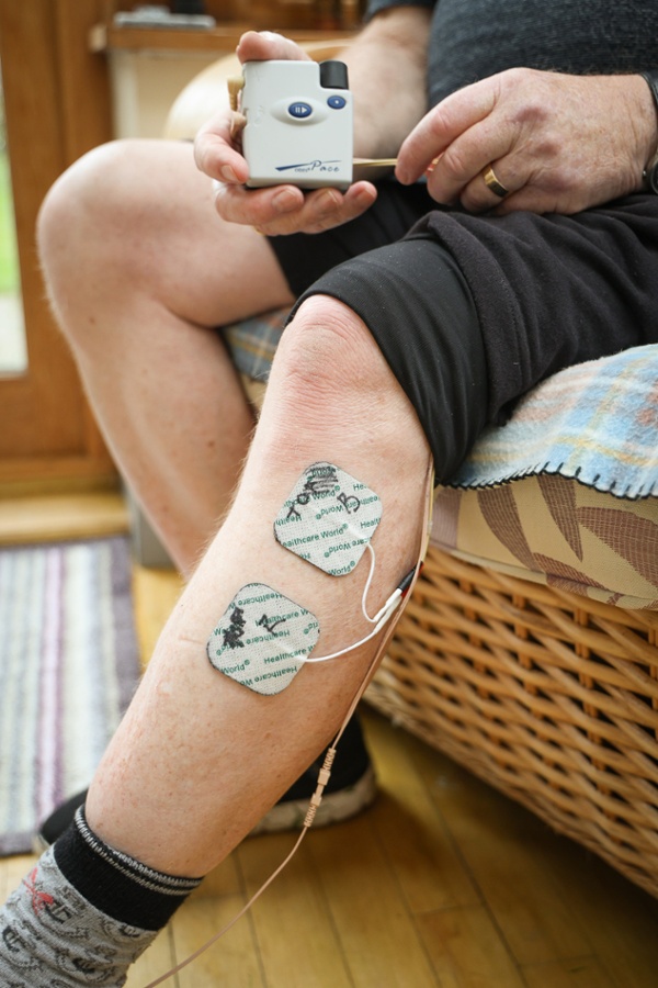 A close-up of a person's lower leg with two sticky electrode pads attached and connected by a wire to a small handheld ODFS Pace device being held in the hand.