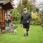 Malcolm Sims, a man in his seventies wearing a black puffer jacket, shorts and a cap, walks across a garden lawn with a small dog on a lead. A black device is visible on his lower leg.