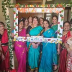 A group of women in colourful traditional Indian saris pose together inside a decorative photo frame reading "Mela 2024", adorned with fabric flowers and greenery, with a sign for the Indian Society of South West Wales visible in the centre.