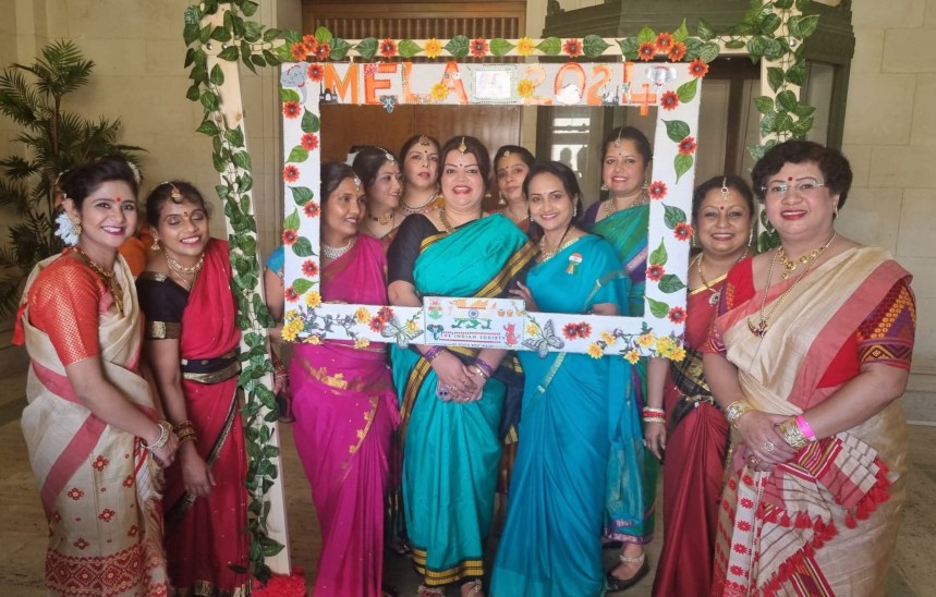 A group of women in colourful traditional Indian saris pose together inside a decorative photo frame reading "Mela 2024", adorned with fabric flowers and greenery, with a sign for the Indian Society of South West Wales visible in the centre.