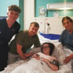 Midwife Michael Nixon in navy scrubs stands on the left alongside Liam John leaning over Shania Jenkins who is lying in a hospital bed, with midwife Leanne Daniel in navy scrubs on the right, all smiling at the camera in a hospital room.