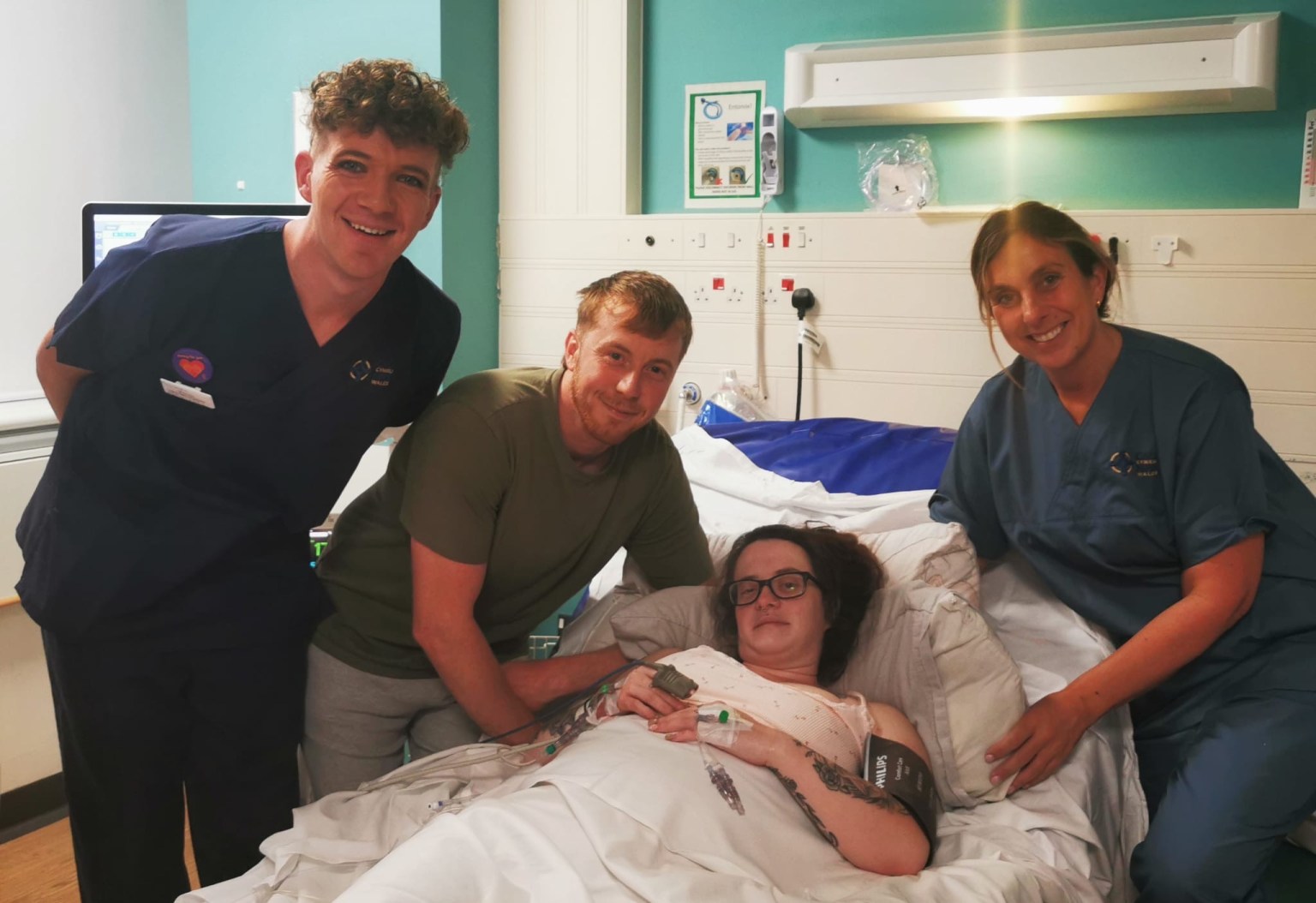 Midwife Michael Nixon in navy scrubs stands on the left alongside Liam John leaning over Shania Jenkins who is lying in a hospital bed, with midwife Leanne Daniel in navy scrubs on the right, all smiling at the camera in a hospital room.