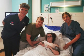 Midwife Michael Nixon in navy scrubs stands on the left alongside Liam John leaning over Shania Jenkins who is lying in a hospital bed, with midwife Leanne Daniel in navy scrubs on the right, all smiling at the camera in a hospital room.