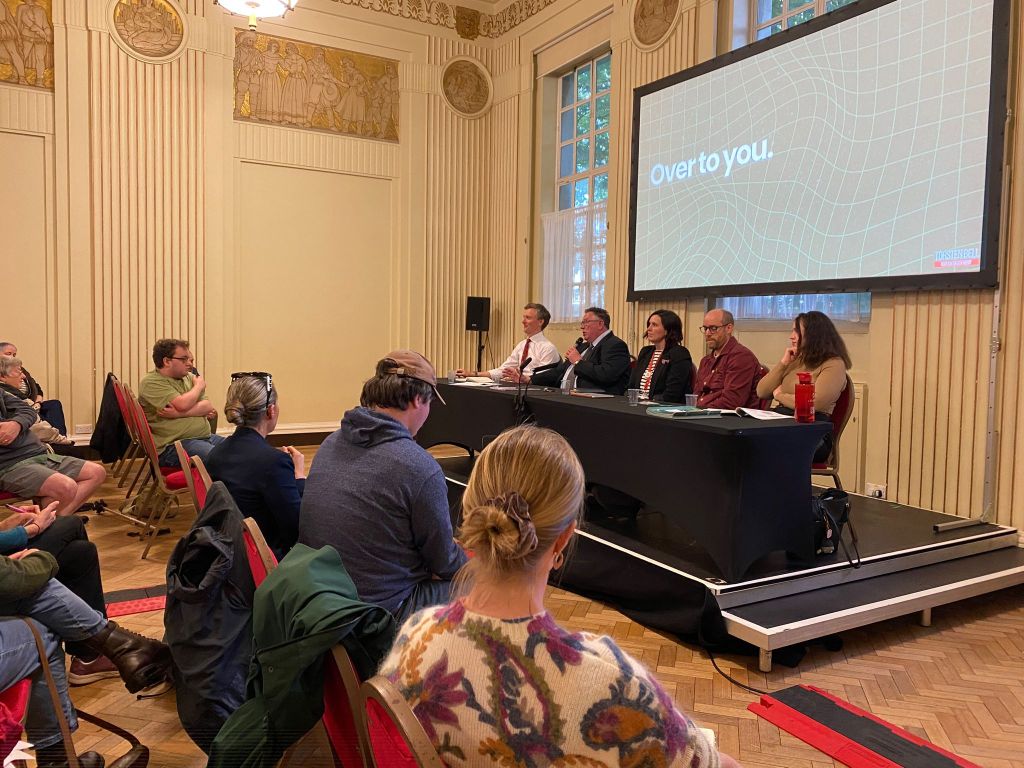 A public meeting in progress in a grand hall, with five panellists seated at a table on a stage and a screen reading "Over to you", with audience members visible on both sides of the room.