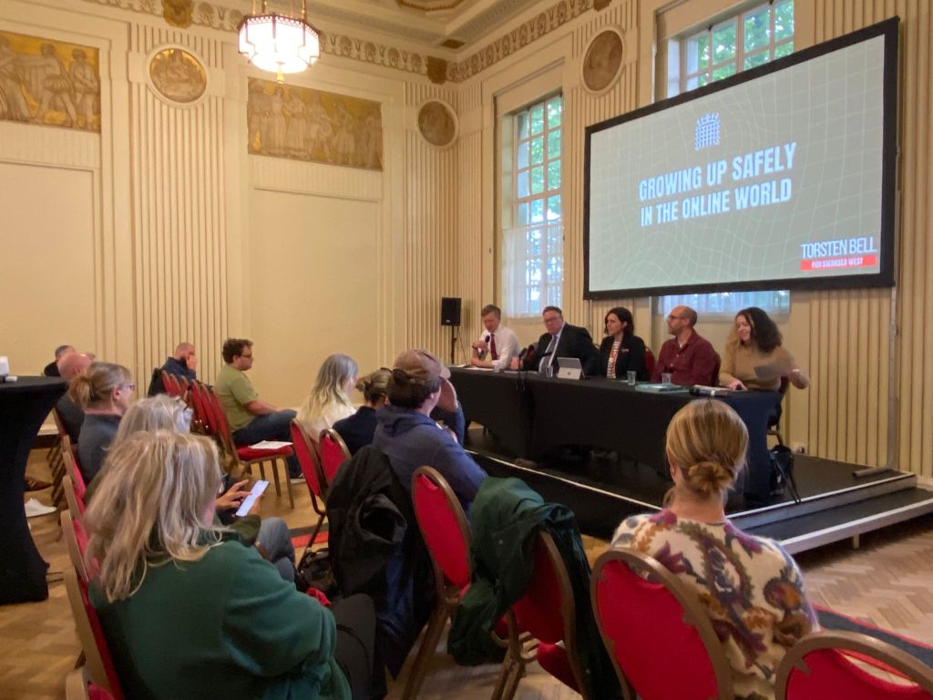 A wide shot of a public meeting in an ornate hall, with an expert panel of five people seated at a table on a raised stage under a large screen reading "Growing Up Safely in the Online World", with rows of audience members in red chairs facing them.