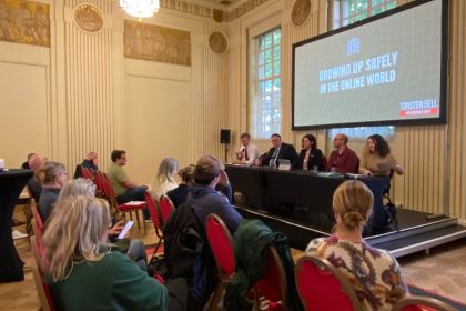 A wide shot of a public meeting in an ornate hall, with an expert panel of five people seated at a table on a raised stage under a large screen reading "Growing Up Safely in the Online World", with rows of audience members in red chairs facing them.