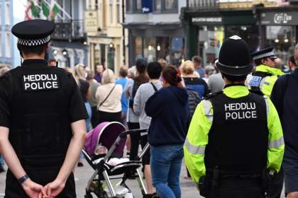Two Dyfed-Powys Police officers with Heddlu/Police markings on their uniforms patrol a busy pedestrianised street in Tenby, with crowds of visitors visible ahead of them and shops in the background