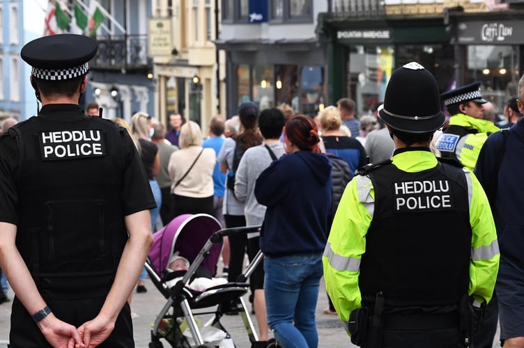 Two Dyfed-Powys Police officers with Heddlu/Police markings on their uniforms patrol a busy pedestrianised street in Tenby, with crowds of visitors visible ahead of them and shops in the background