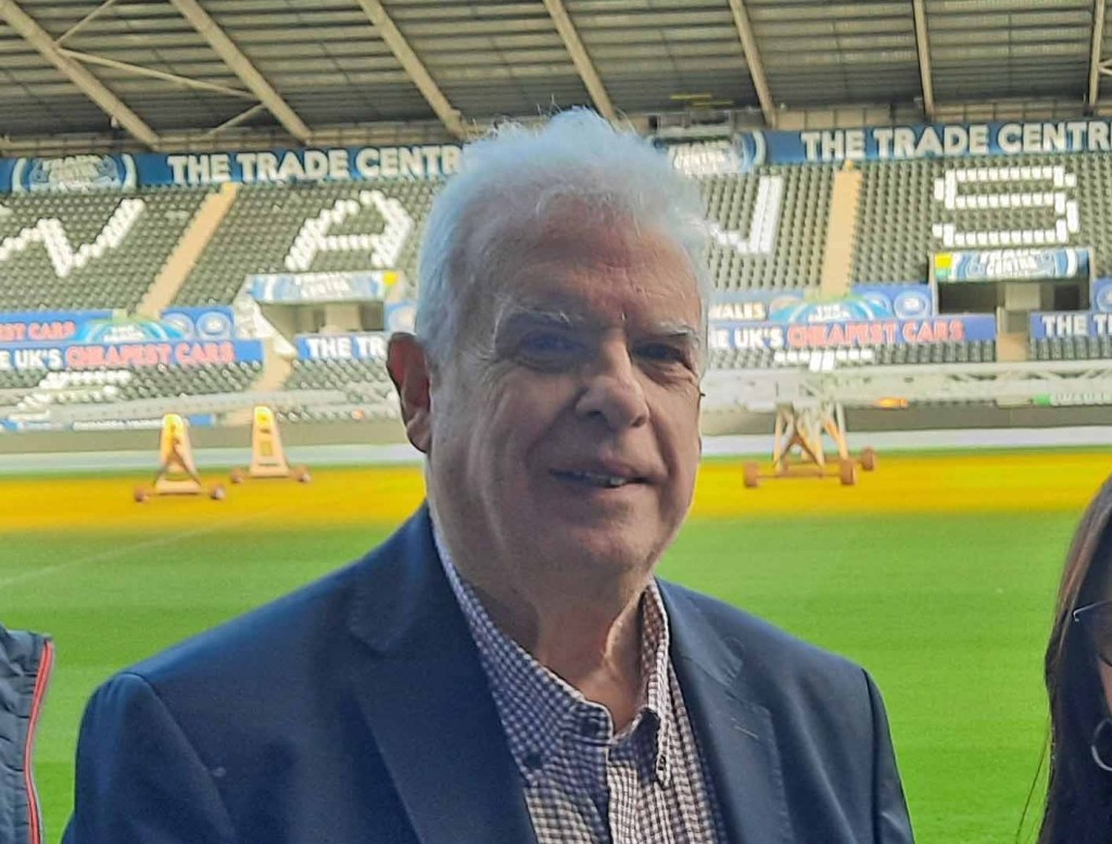 Cllr Robert Francis-Davies, a white-haired man in a navy blazer and checked shirt, smiling on the pitch at the Swansea.com Stadium with the stands and advertising hoardings visible in the background.