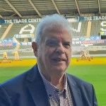 Cllr Robert Francis-Davies, a white-haired man in a navy blazer and checked shirt, smiling on the pitch at the Swansea.com Stadium with the stands and advertising hoardings visible in the background.