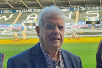 Cllr Robert Francis-Davies, a white-haired man in a navy blazer and checked shirt, smiling on the pitch at the Swansea.com Stadium with the stands and advertising hoardings visible in the background.
