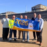 Six men stand on Aberavon Beach holding a large blue and yellow 2026 Seaside Award flag, with the beach promenade, Beach Rescue station and blue sky visible behind them.