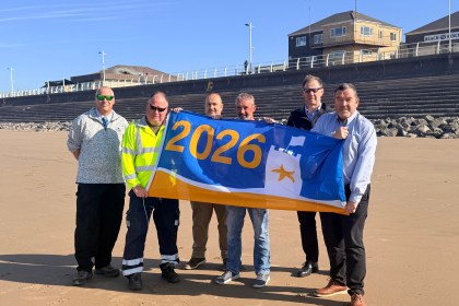 Six men stand on Aberavon Beach holding a large blue and yellow 2026 Seaside Award flag, with the beach promenade, Beach Rescue station and blue sky visible behind them.