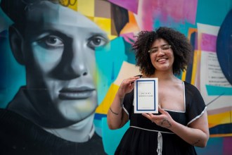 Sasha Debevec-McKenney smiles as she holds her poetry collection Joy Is My Middle Name in front of a colourful mural featuring a black and white portrait, wearing a black dress and clear-framed glasses with curly hair.