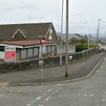 A street view of the former Ysgol Gymraeg Tan Y Lan school site in Morriston, showing the Victorian stone building with its distinctive gable end and arched window behind a single-storey timber-clad extension, with a Lambert Smith Hampton "For Sale" board mounted on the wall facing the road and a no-entry sign in the foreground