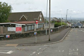 A street view of the former Ysgol Gymraeg Tan Y Lan school site in Morriston, showing the Victorian stone building with its distinctive gable end and arched window behind a single-storey timber-clad extension, with a Lambert Smith Hampton "For Sale" board mounted on the wall facing the road and a no-entry sign in the foreground