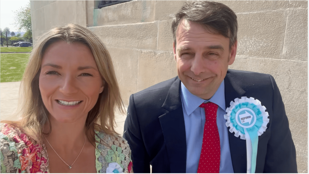 Newly-elected Reform UK Senedd Members Francesca O'Brien and Steven Rodaway pictured outside the count venue — Francesca on the left with long blonde hair wearing a multicoloured crochet cardigan, and Steven on the right in a navy suit, light blue shirt, red polka-dot tie and Reform UK rosette