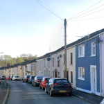 A residential street in Felinfoel showing a row of terraced houses in pale colours with cars parked along the roadside under a blue sky.