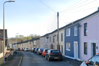 A residential street in Felinfoel showing a row of terraced houses in pale colours with cars parked along the roadside under a blue sky.