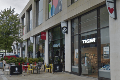Exterior of the Flying Tiger store in Swansea on Princess Way, with the brand's red heart and toy soldier logo on a sign jutting out from the building. Coffee#1 and Zinco Lounge sit alongside, with outdoor seating in the foreground.