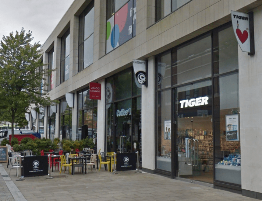 Exterior of the Flying Tiger store in Swansea on Princess Way, with the brand's red heart and toy soldier logo on a sign jutting out from the building. Coffee#1 and Zinco Lounge sit alongside, with outdoor seating in the foreground.