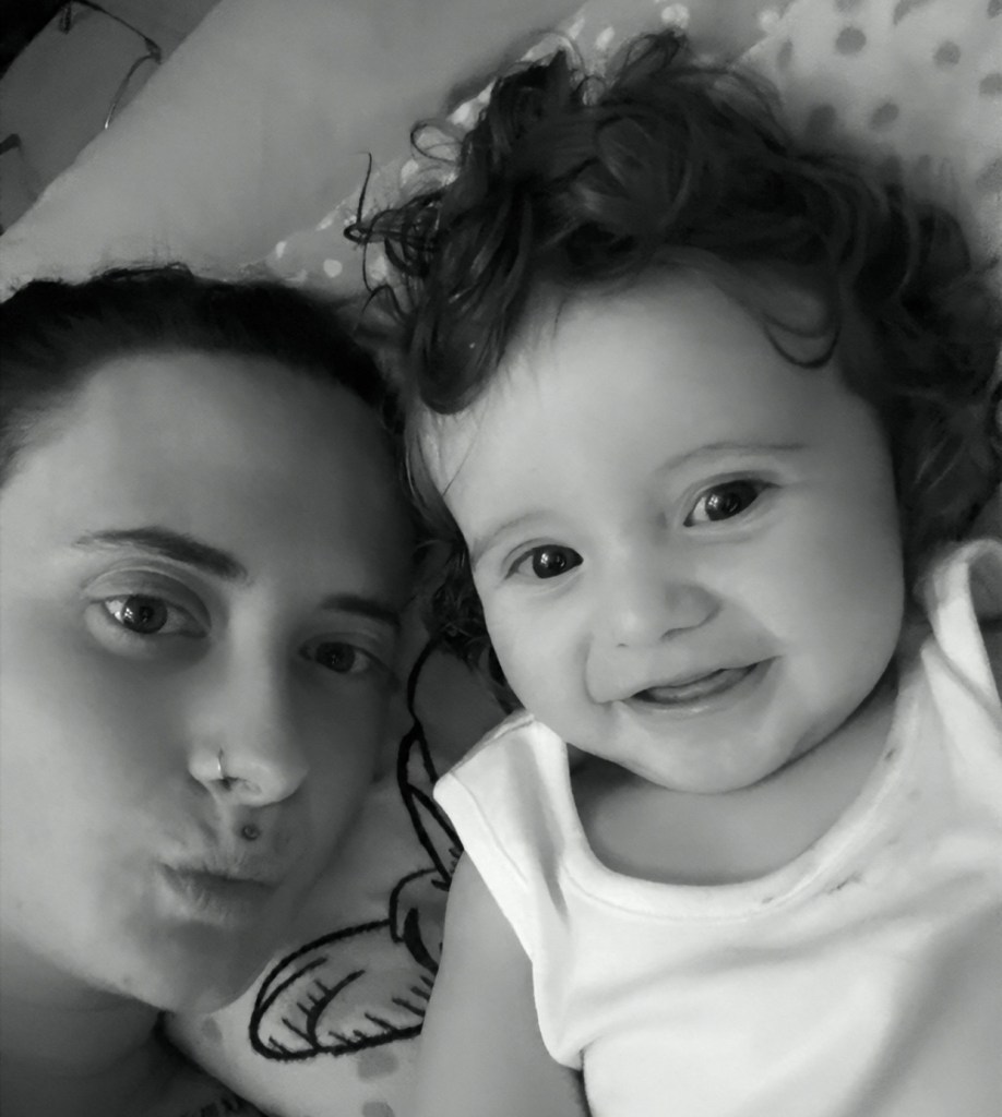 A black and white close-up photograph of Shania Jenkins lying alongside her smiling baby Tobi, who has dark curly hair and is wearing a white babygrow.
