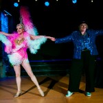 Two performers on a stage — one in a full pink feathered showgirl costume with a tall headdress, the other in a blue sequinned jacket — holding hands with arms outstretched under colourful stage lighting.