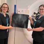 Two NHS staff members in black uniforms hold up a large X-ray film in front of a radiotherapy CT scanner at the South West Wales Cancer Centre in Singleton Hospital, Swansea