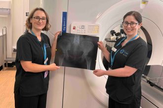 Two NHS staff members in black uniforms hold up a large X-ray film in front of a radiotherapy CT scanner at the South West Wales Cancer Centre in Singleton Hospital, Swansea