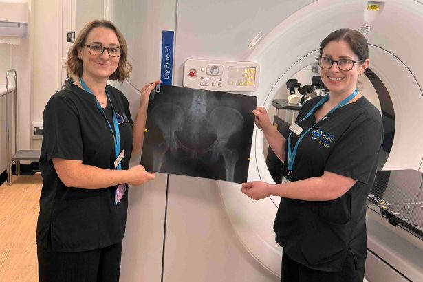 Two NHS staff members in black uniforms hold up a large X-ray film in front of a radiotherapy CT scanner at the South West Wales Cancer Centre in Singleton Hospital, Swansea